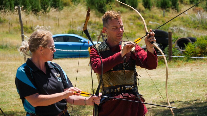 A man dressed in armour instructing a woman in archery.
