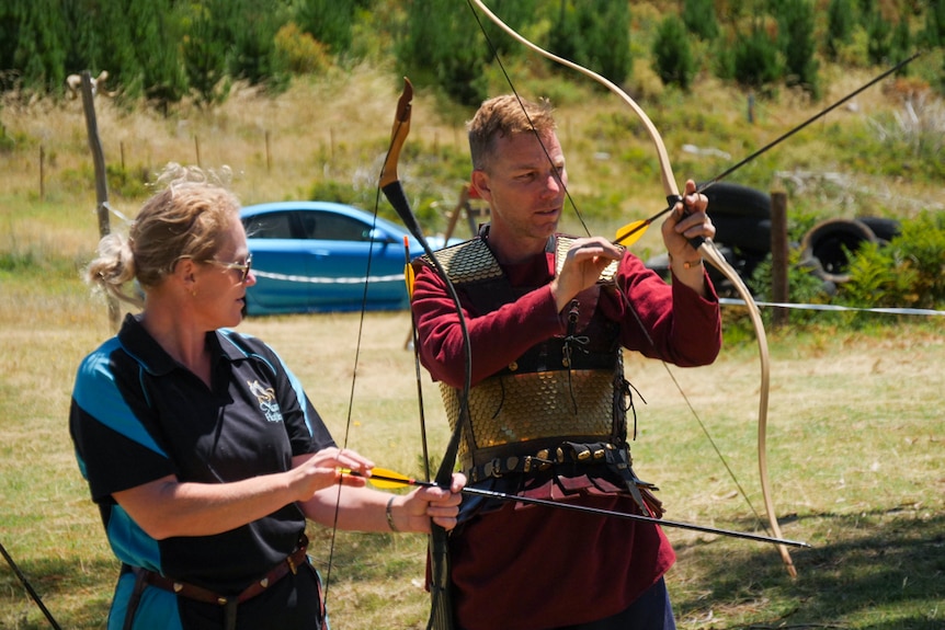 A man dressed in armour instructing a woman in archery.