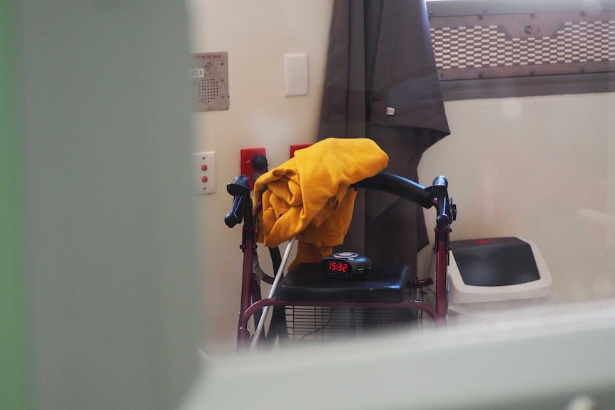 A walking frame with an alarm clock placed on the seat and a yellow jumper folder over it seen through a prison window.
