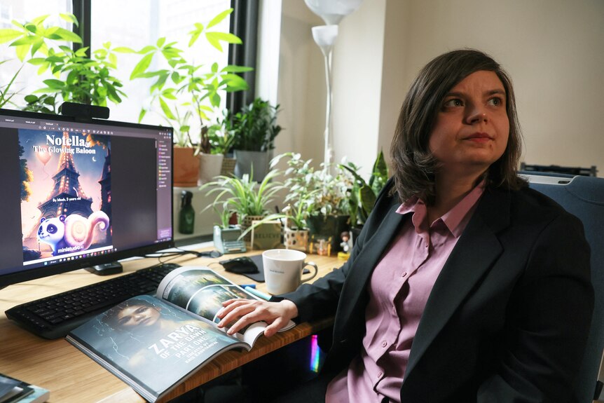 A woman with shoulder-length hair, wearing a suit jacket and a pink shirt, sits in front of a computer displaying a book cover.