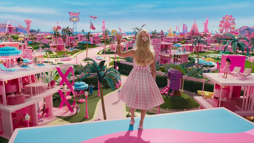 The actor Margot Robbie in a pink dress and long blonde hair playing the character Barbie in the movie, standing over barbieland