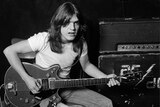 A young Malcolm Young holds a guitar as he poses, sitting on an amp