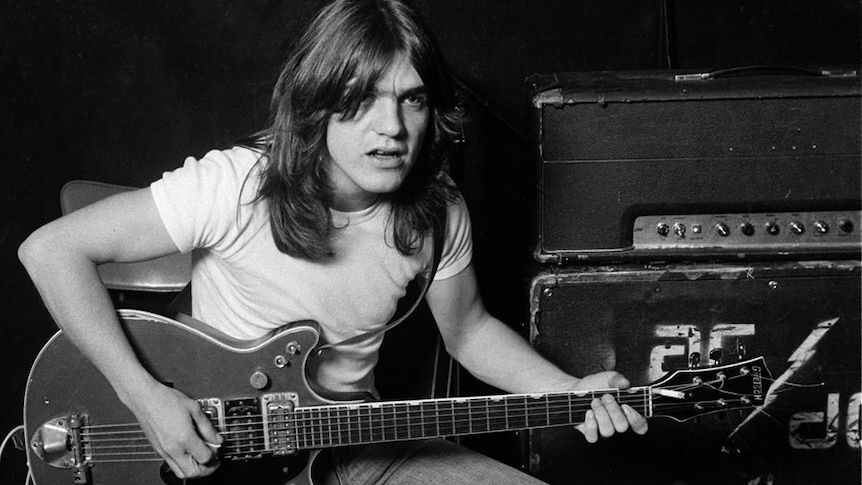 A young Malcolm Young holds a guitar as he poses, sitting on an amp