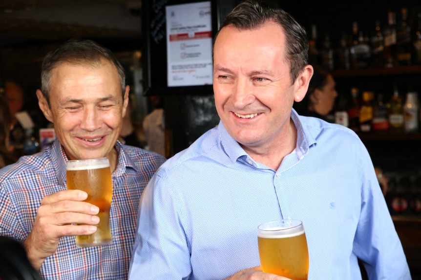 Two men wearing shirts stand drinking pints of beer in a pub.