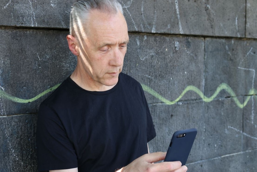 Before a backdrop of water, a man stands against a green railing in black t-shirt, with arms folded, looking forward.