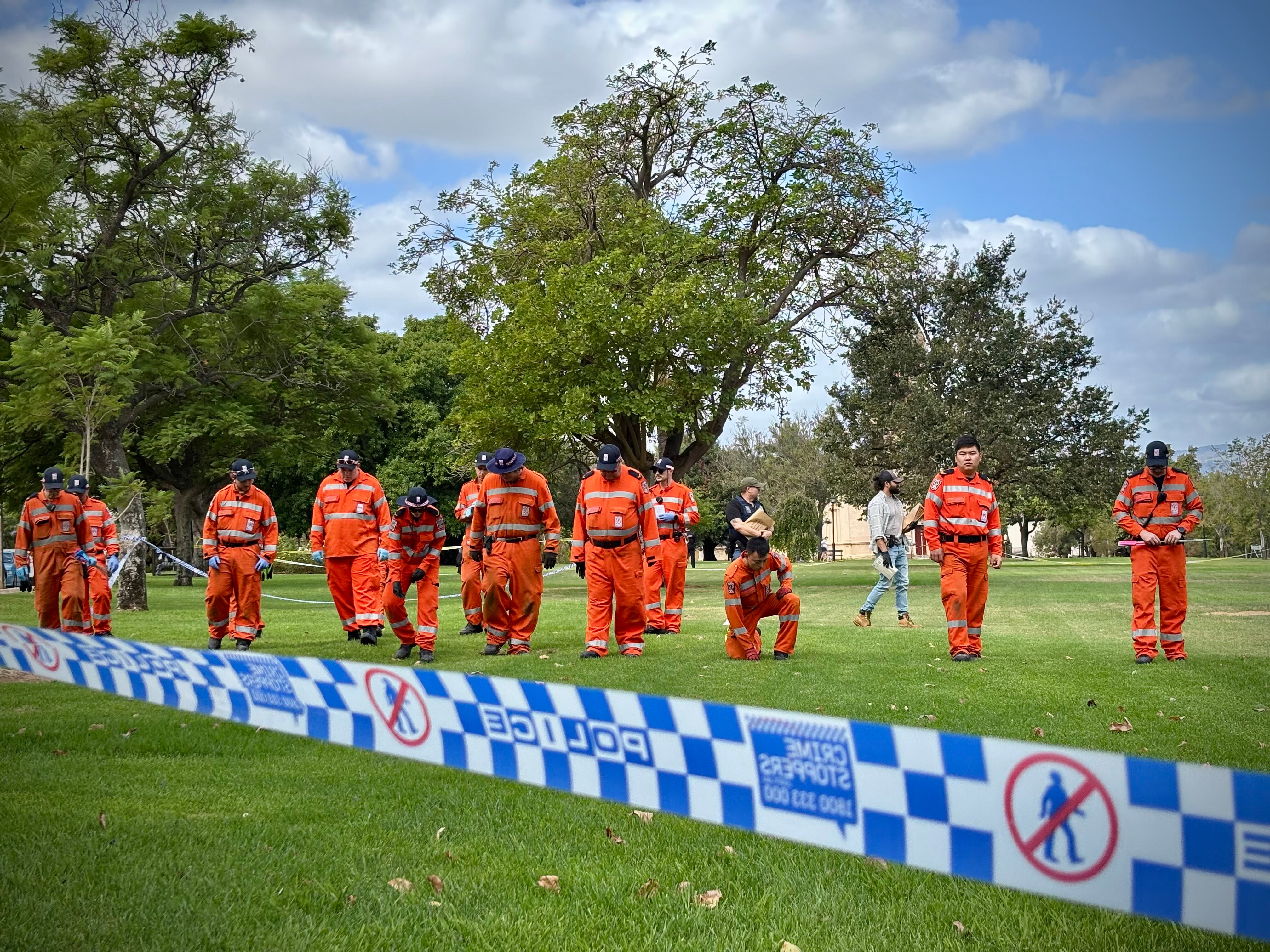 A group of SES volunteers in orange uniforms walk in a line across a park behind crime scene tape