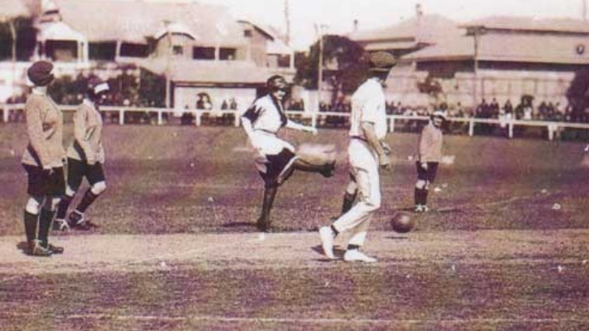 A female football player kicks a ball watched by other players and spectators