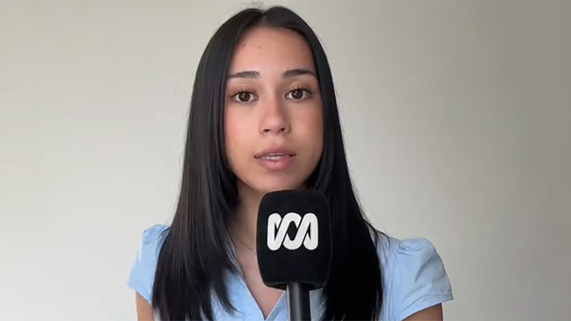 Young woam with dark shoulder length hair wearing a light blue shirt. She is holding an ABC microphone.