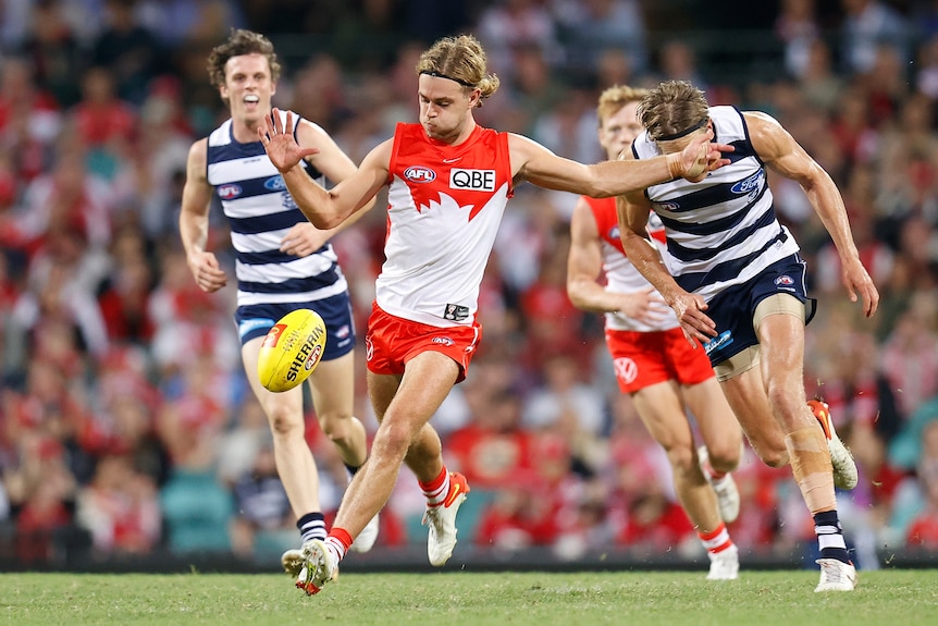 A Sydney Swans player looks down as he kicks the football on the run while Geelong players trail behind him.