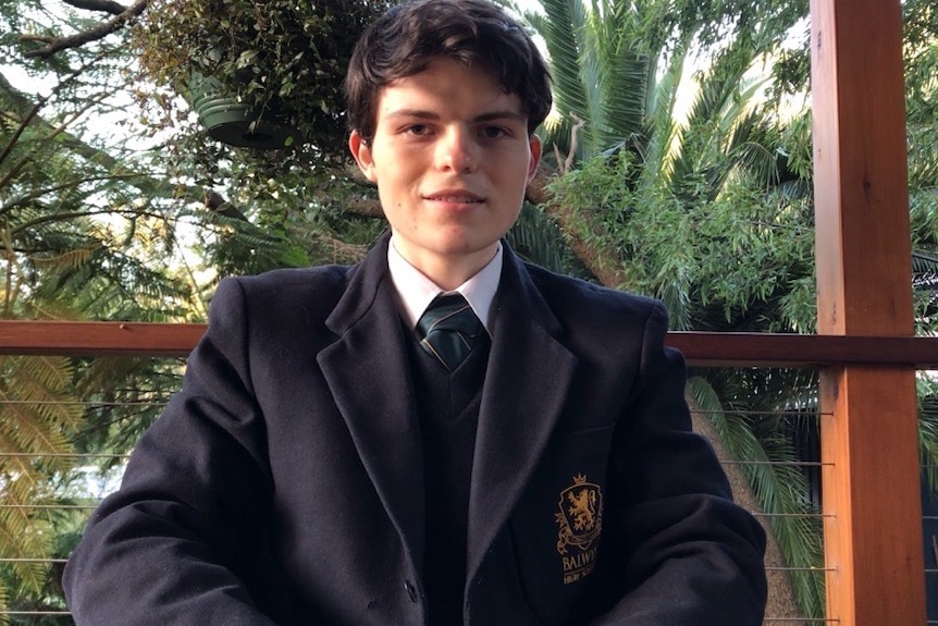Liam Cruise wearing the Balwyn High School uniform with jacket and tie, sitting in front of some lush green trees.
