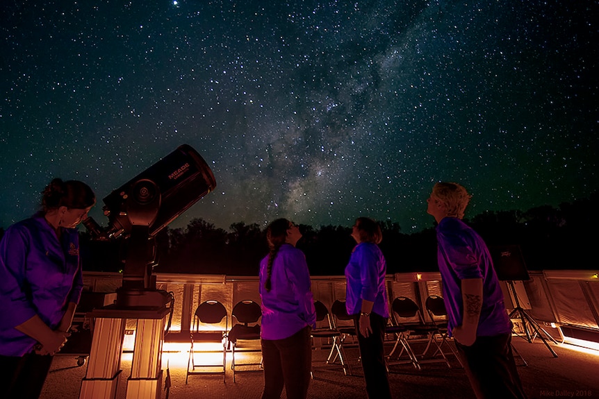 Four people stand under a starry night's sky in the outback. One looks through a telescope.