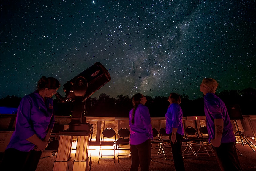 Four people stand under a starry night's sky in the outback. One looks through a telescope.