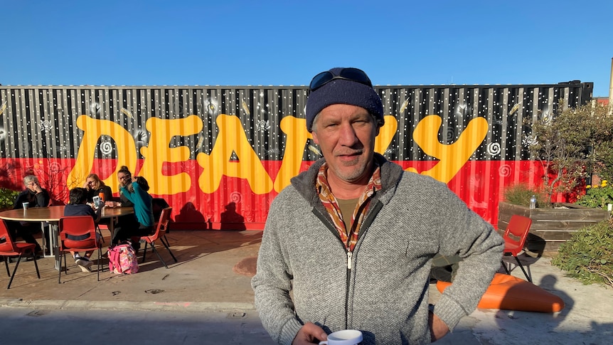 Man standing in front of a shipping container painted with the Aboriginal Flag and the word 'Deadly'