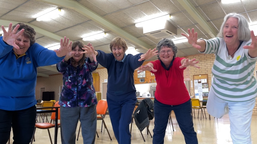 A group of five women standing side by side, smiling with their arms out 