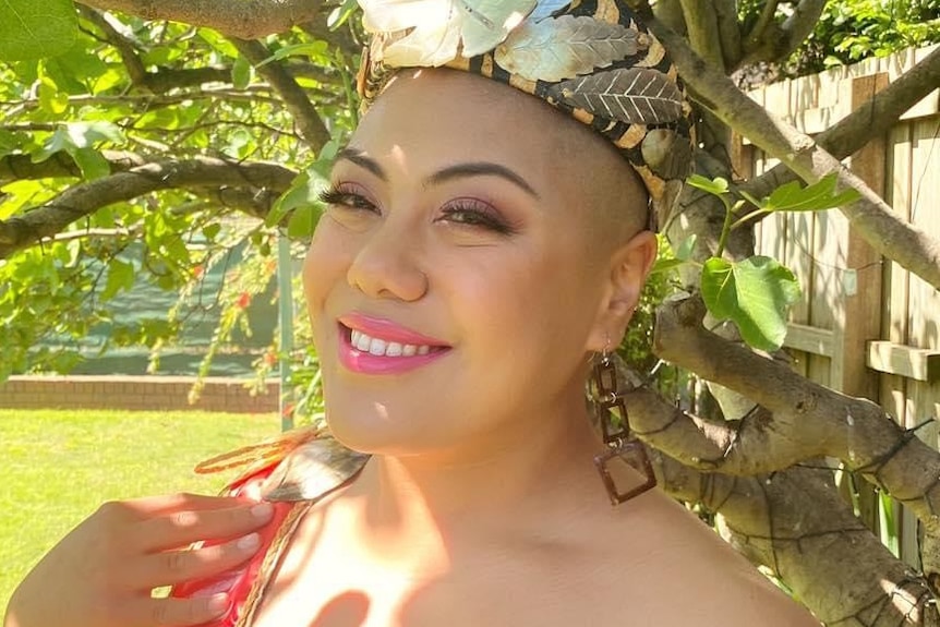 Woman stands under tree and smiles to camera, wearing a women floral headband and pink dress.