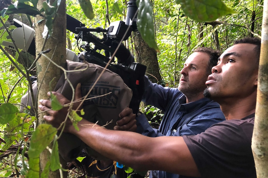 Nelson holding camera standing next to Yockno in thick green jungle looking up.