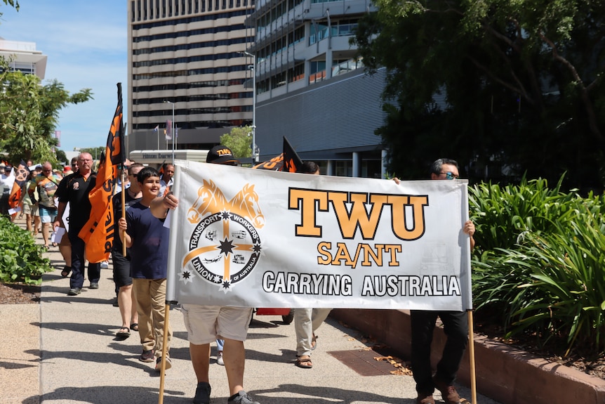 A group of protesters marching down a footpath as someone at the front holds up a large sign, on a sunny day.