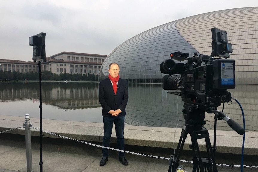 A man in a suit and red scarf is filmed by a camera