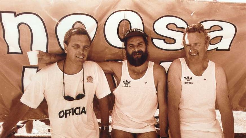 A sepia photo of three men sitting in front of a banner saying Noosa, sitting smiling in t-shirts and togs.