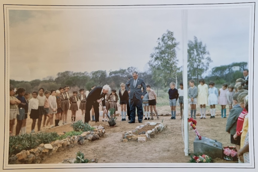 A man in a suit with a shovel leans over planting a small sapling as others look in. 