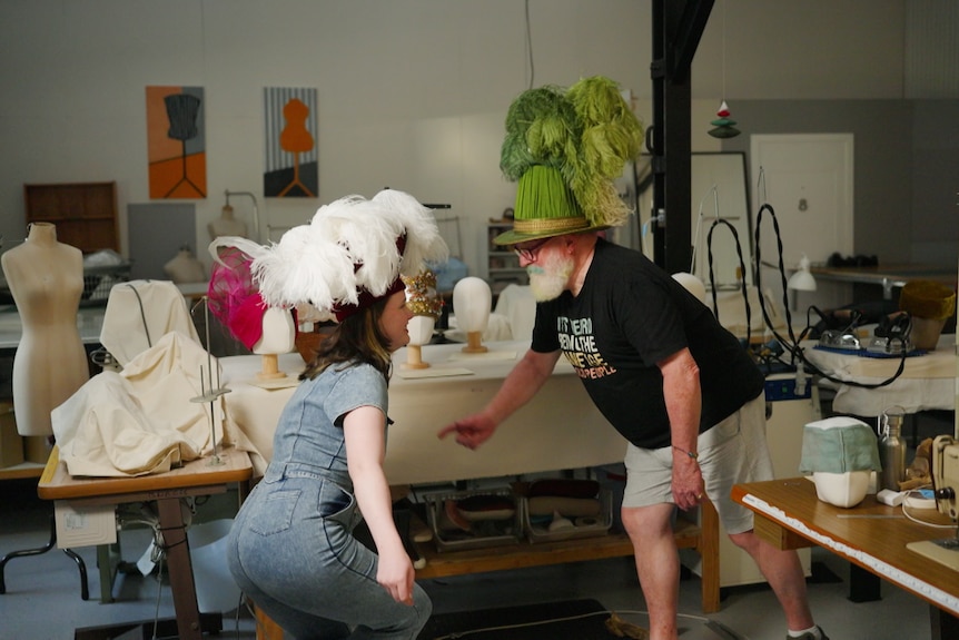 Kirrilly, left, wears a jean jumpsuit and a white feather hat and bobs down mid-dance with Rick, right, wearing a green hat.