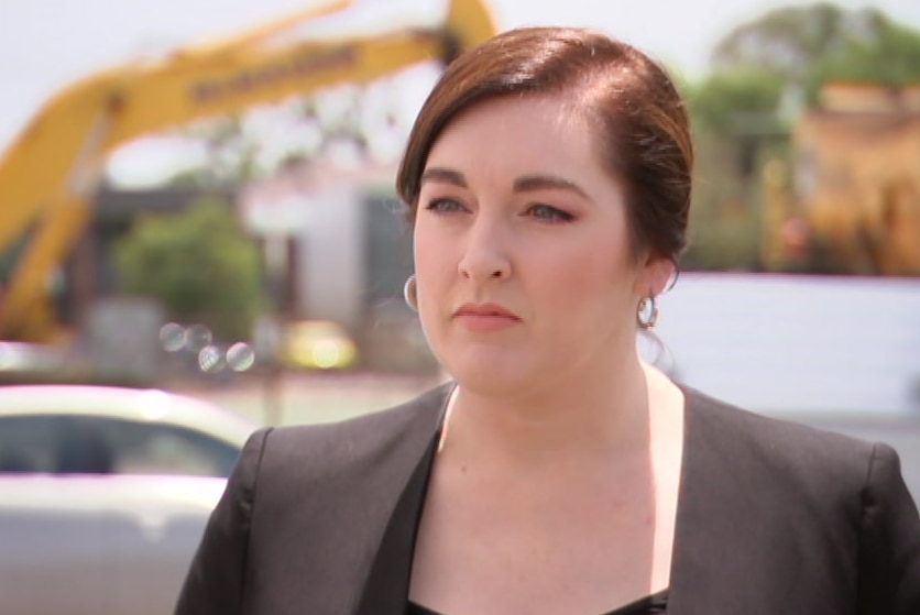 A woman with brown hair, wearing a dark suit, looking stern.