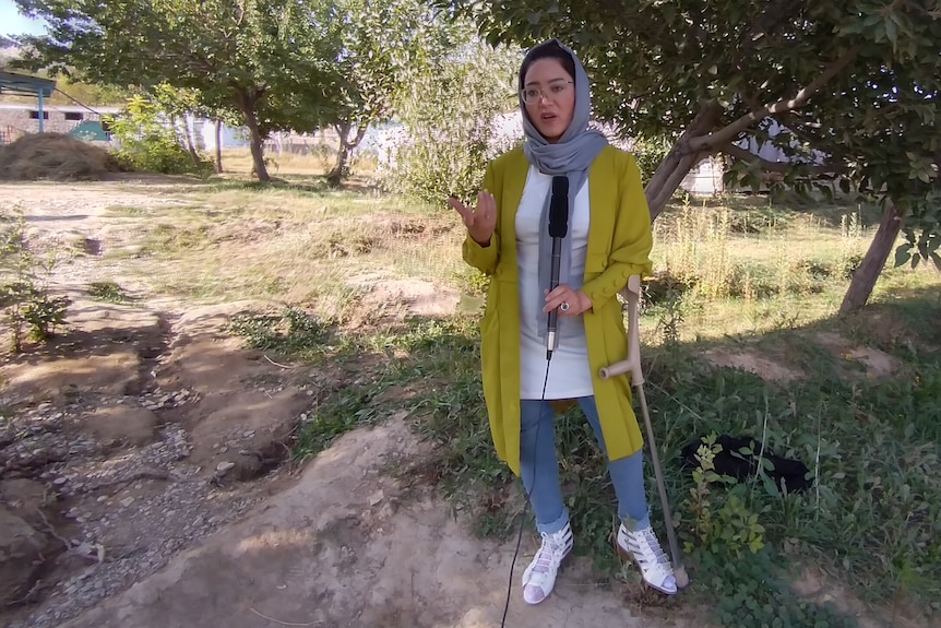 A woman stands near trees holding a walking stick.