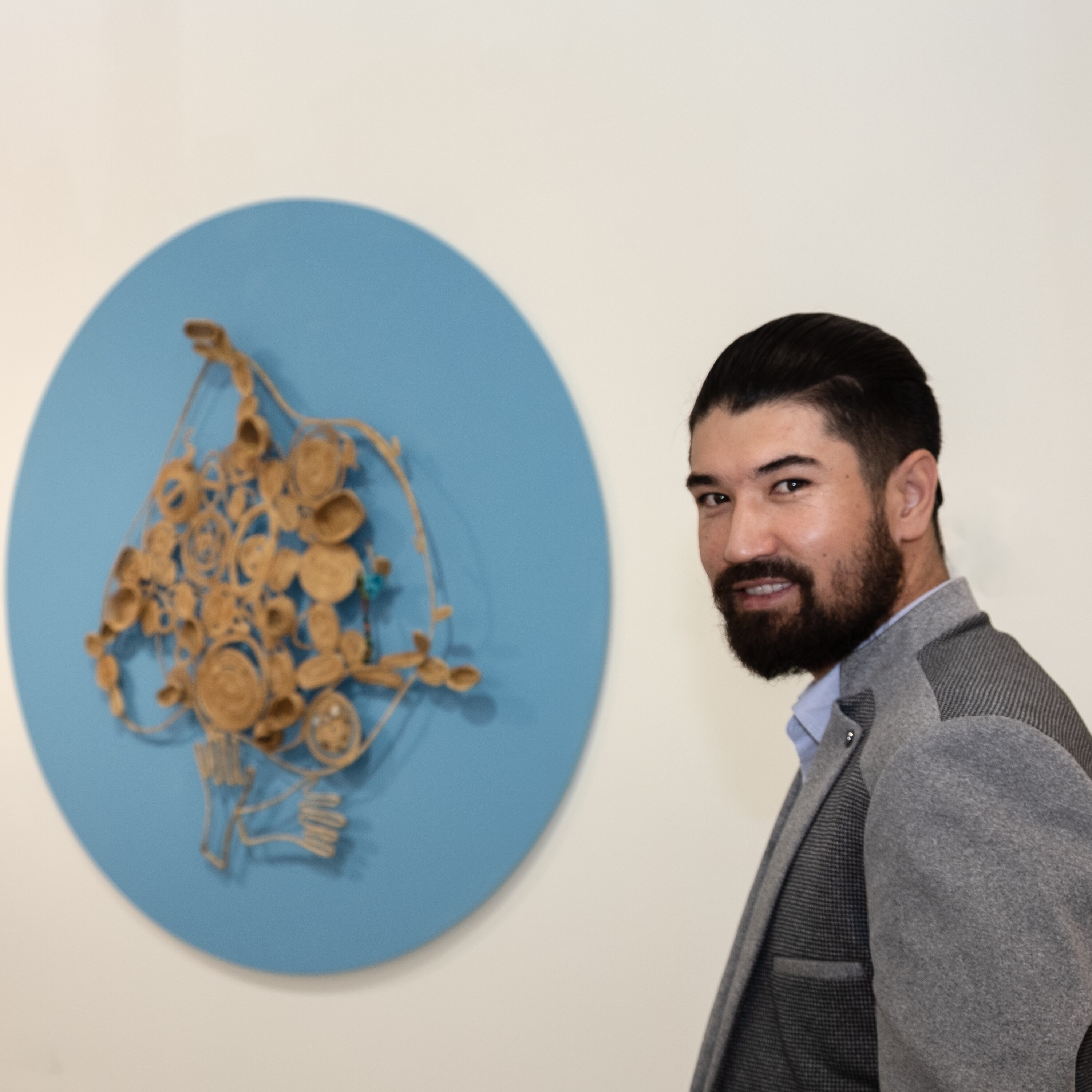 A man with dark hair and a beard stands beside an artwork hanging on a wall.