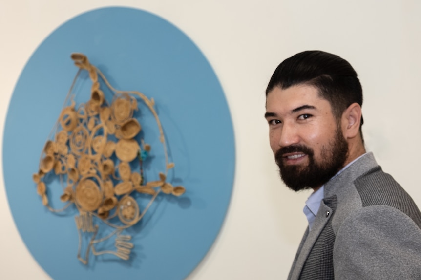 A man with dark hair and a beard stands beside an artwork hanging on a wall.