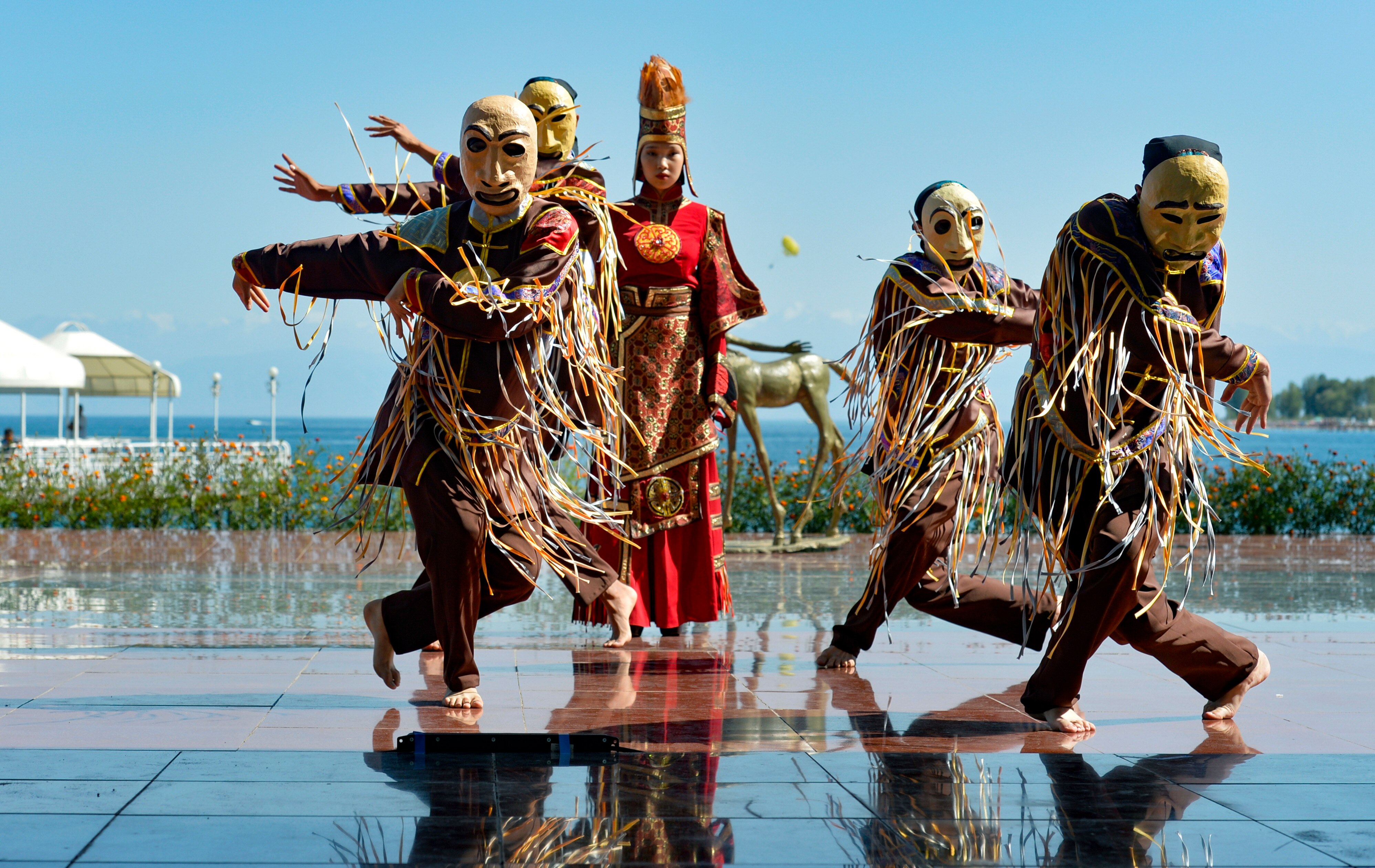 Masked performers dance across tiles.  a garden bed and the ocean are in the distant background. 