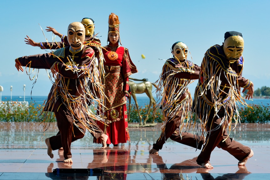 Masked performers dance across tiles. a garden bed and the ocean are in the distant background. 