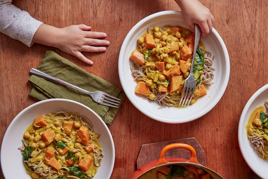 Three bowls of chickpea sweet potato curry on a set table, with a child's hand reaching for a fork.