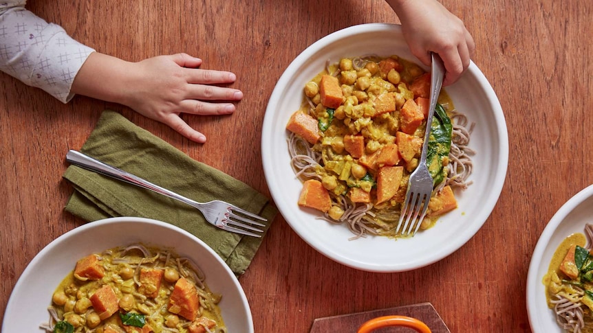 Three bowls of chickpea sweet potato curry on a set table, with a child's hand reaching for a fork.