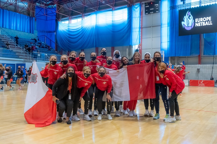 Malta's netball team adn officials standing on court wearing red jumpers with the Maltese cross and holding maltese flags