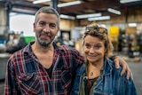 A man in a plaid shirt with a greying beard puts his arm around his wife who is blonde and wearing a denim jacket