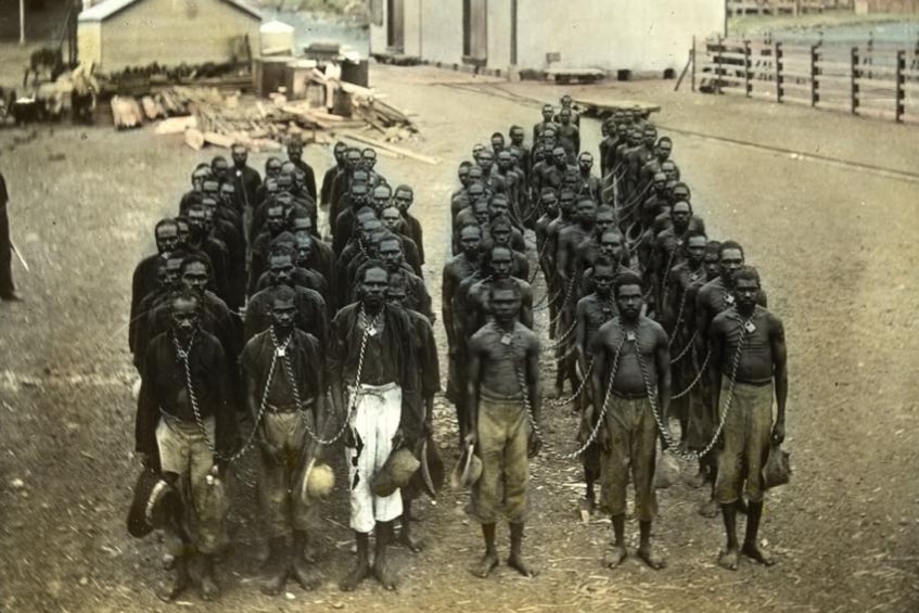Dozens of Aboriginal men stand chained together in a dirt area with dilapidated buildings behind them.
