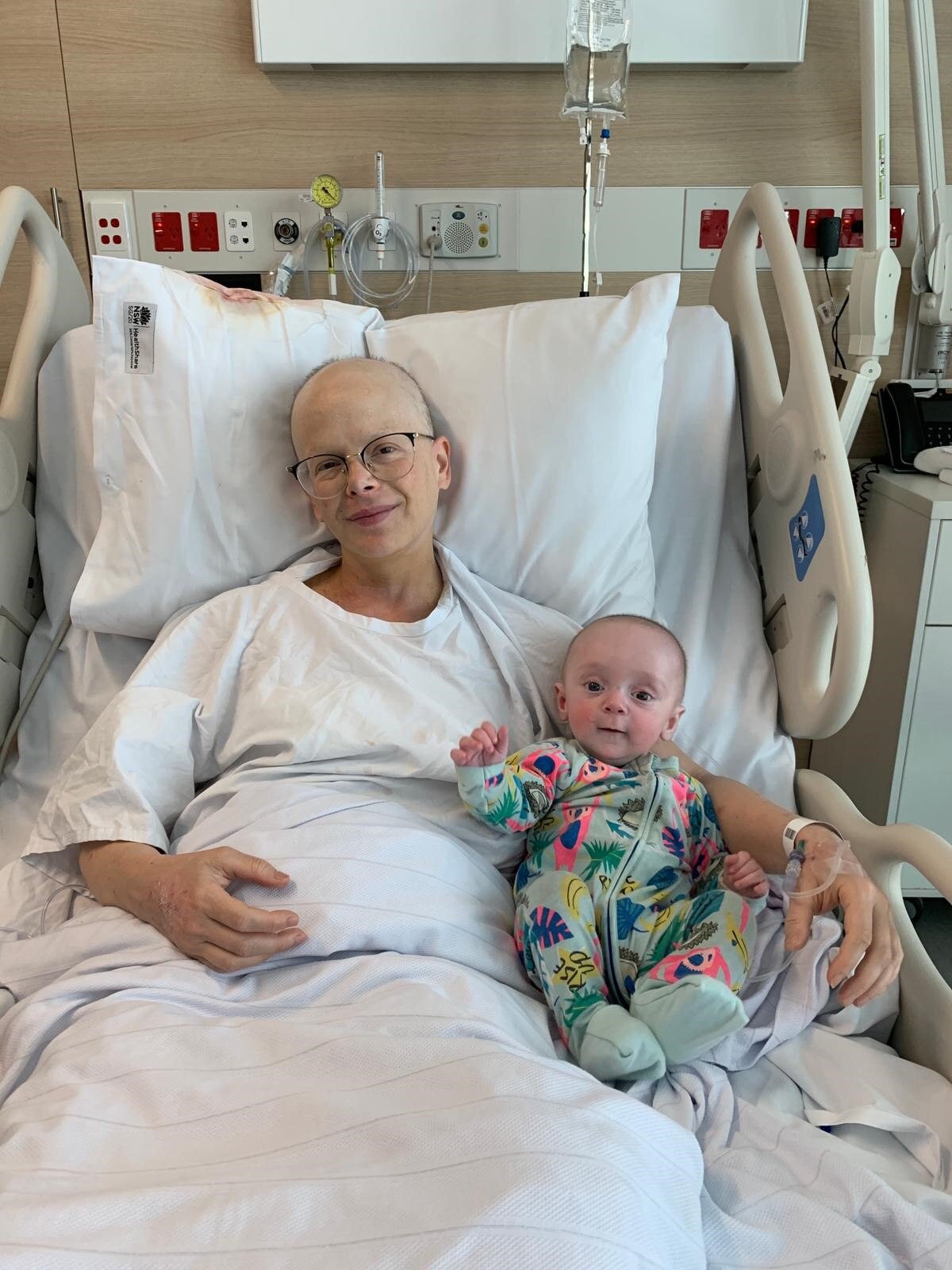 A woman with a bald head and glasses lying in a hospital bed, snuggled up to a baby in a brightly coloured onesie