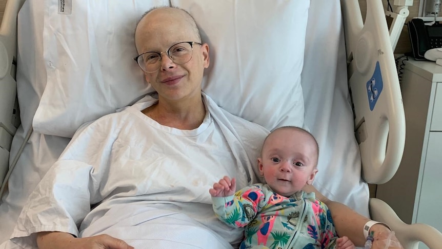 A woman with a bald head and glasses lying in a hospital bed, snuggled up to a baby in a brightly coloured onesie