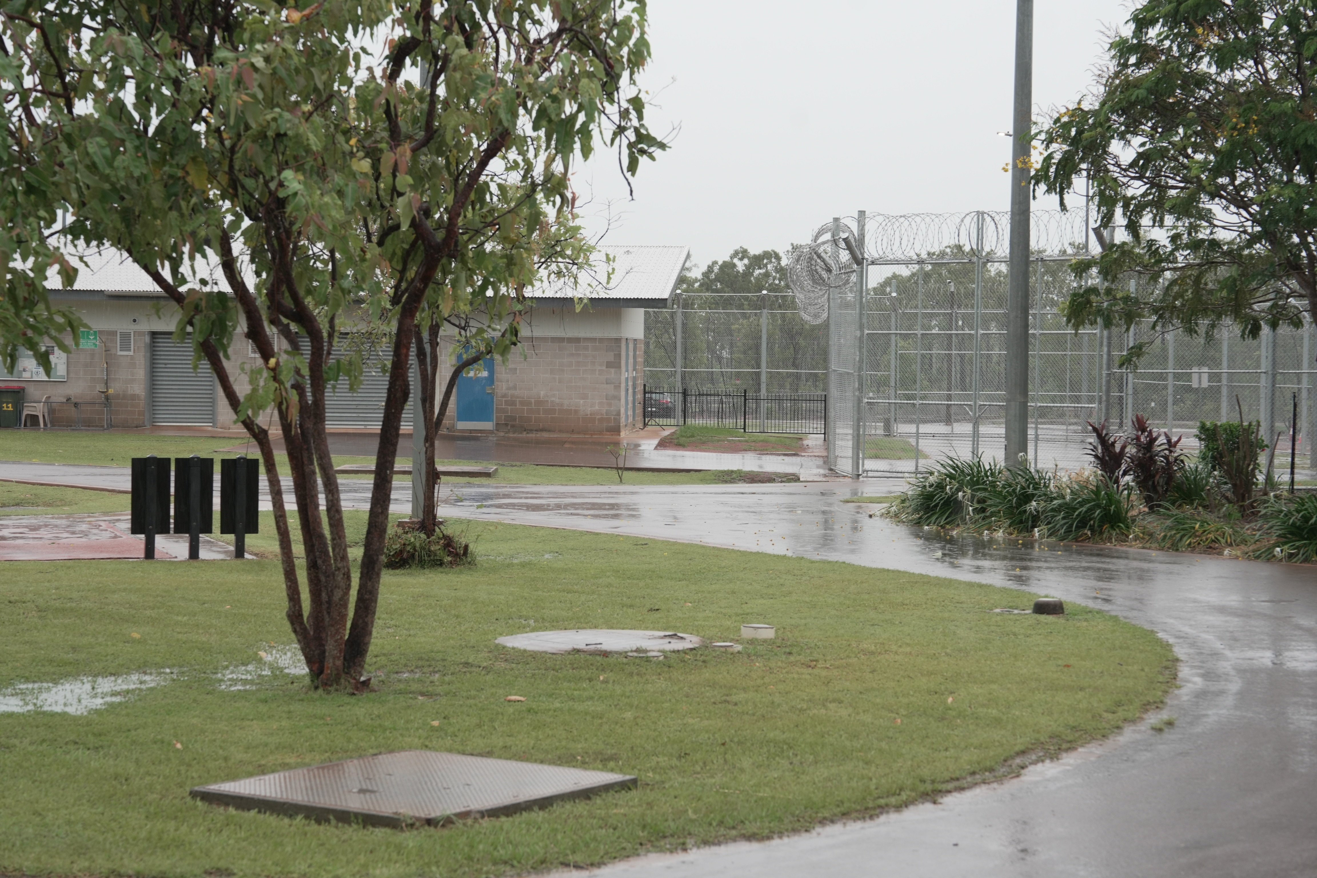 Un plano amplio de hierba verde, un sendero a su alrededor, un edificio de concreto en la distancia, cercas de alambre alrededor, en la distancia.