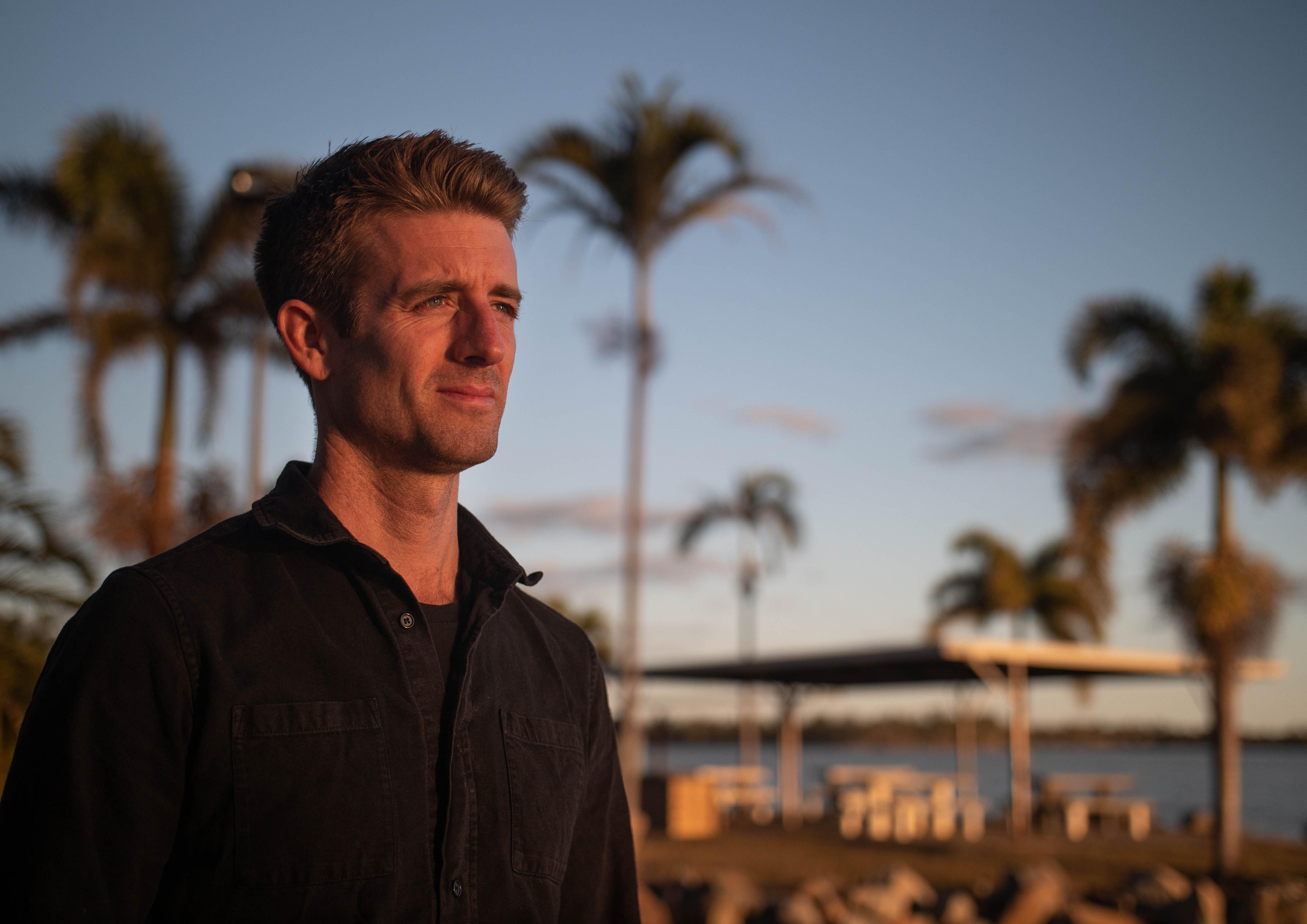 A man wearing a black shirt, in front of a lake with palm trees, soft afternoon sun lights his face.