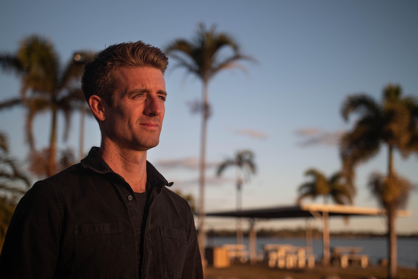 A man wearing a black shirt, in front of a lake with palm trees, soft afternoon sun lights his face.