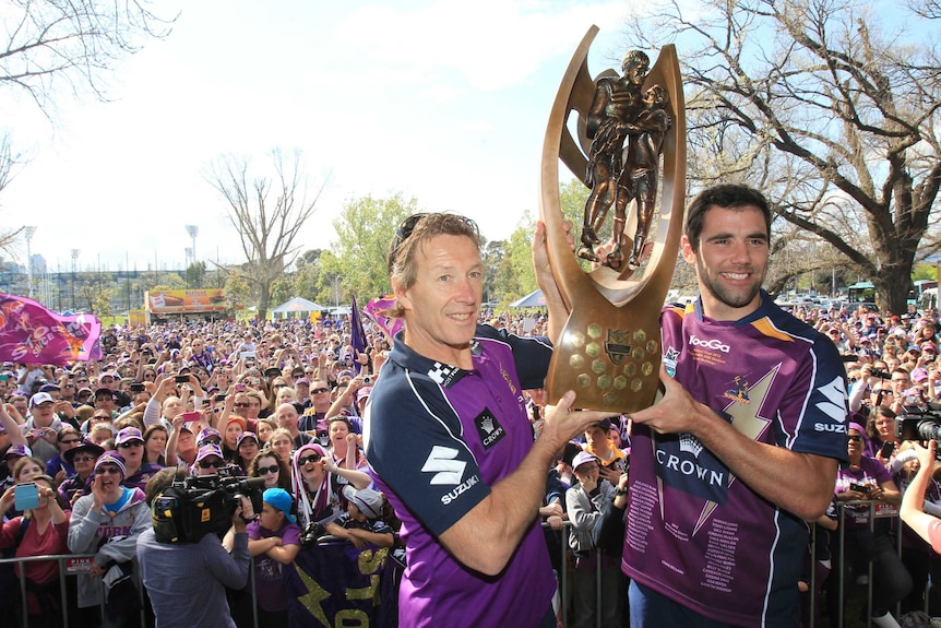 An NRL team coach and captain smile as they hold the trophy, with a big crowd standing behind them.