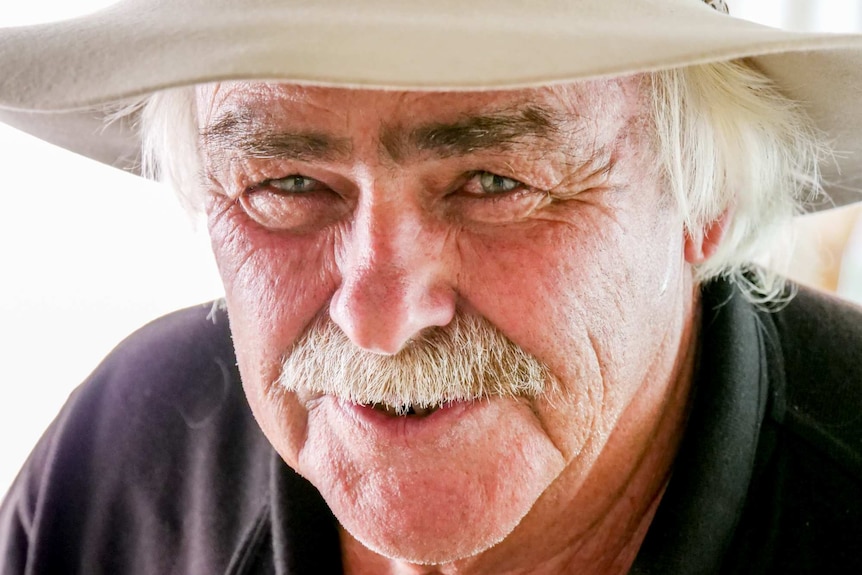 An older man wearing a hat stands smiling up at the camera.