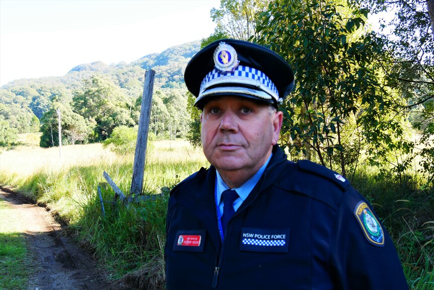 A middle-aged police officer in full uniform standing in a rural area.