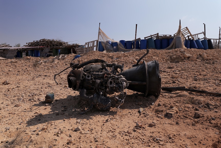 A broken part from a drone on top of sand with water canisters in the background 