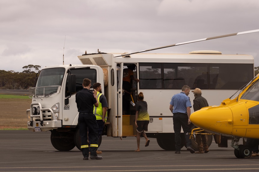 People board a bus, with the yellow nose of a helicopter visible next to it. 