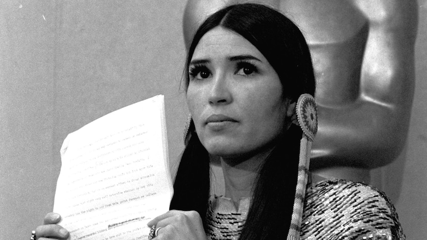 A woman in Native American dress holds up a sheet of paper, with the academy award statue behind her