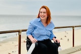 A woman with red short hair in a light blue shirt is in a wheelchair and laughing while in front of a beach outside 