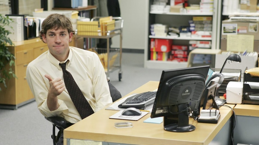 Jim Halpert from The Office sits casually at his desk with his tie loosened giving a thumbs-up