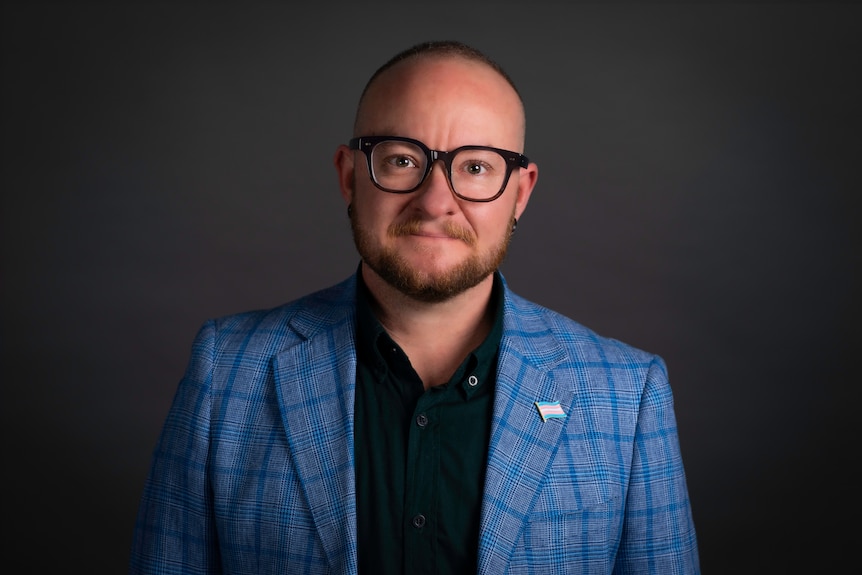 A man wearing a blue suit jacket, a black shirt and thick black glasses